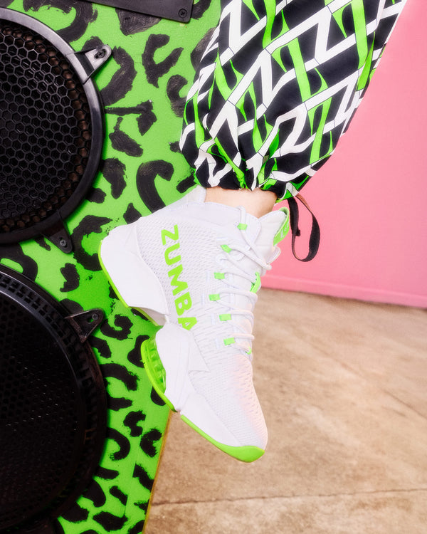 A white Zumba sneakers with neon green accents, worn by a model whose legs are also clad in a black, white, and neon green patterned sweatpants, set against a background with a pink wall and a speaker.