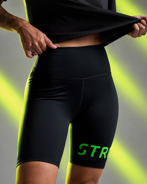 Female model lifting a black top to show the high waistband of the black 'STRONG iD' biker shorts, against a neon light beam background.