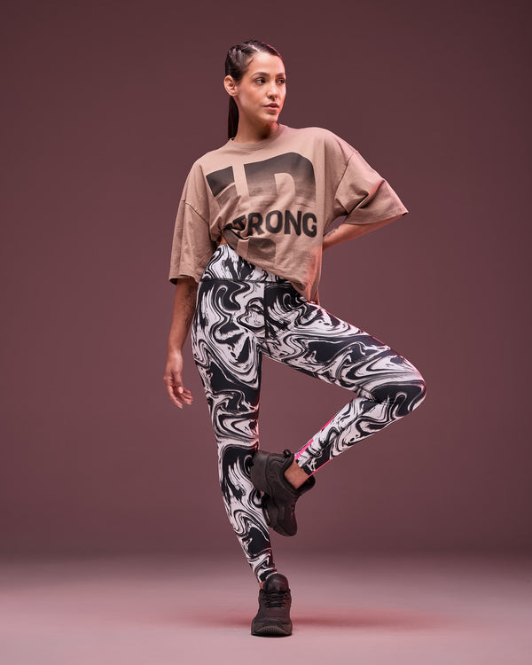 Taupe oversized T-shirt with a large black “STRONG ID” graphic front print, shown full length on a female model wearing black, white, and gray marble-print leggings with pink accents and black sneakers in a dynamic pose against a dark mauve studio background.