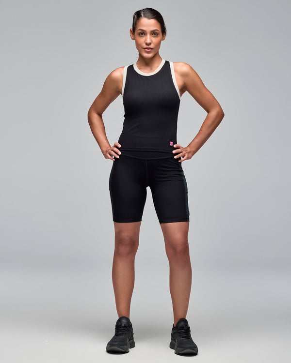 Black sleeveless athletic tank with white trim at the neckline and armholes, shown on a female model with black fitted biker shorts and black sneakers against a light gray studio background.