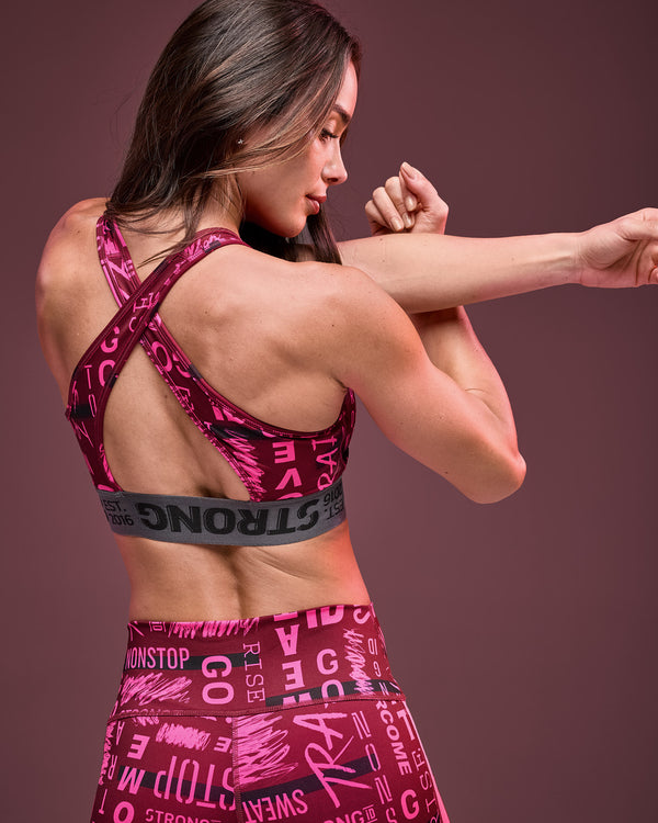 Burgundy and pink graphic-print sports bra with crisscross back straps and a gray elastic under-band, shown from the back on a female model wearing matching high-waisted leggings against a dark mauve studio background.