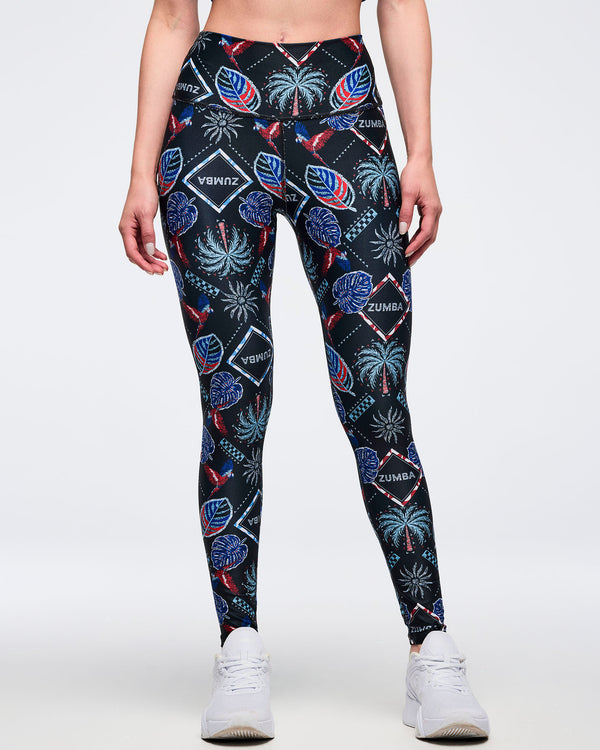 Front view of high-waisted black tropical-print leggings with “ZUMBA” diamond logos and colorful palm-and-parrot pattern; female model wears white sneakers on a white background.