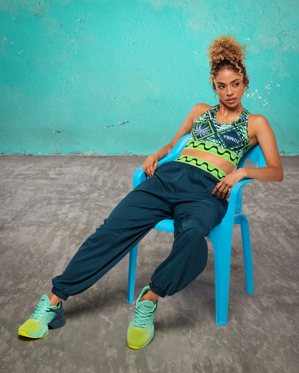 Teal cargo jogger pants with a neon wave print waistband, worn by a female model seated in a blue chair; styled with a green patterned scoop bra and aqua-to-lime sneakers against a teal wall backdrop.