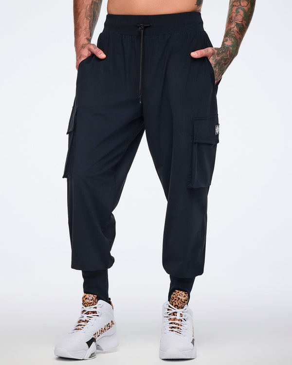 Black cargo jogger pants with oversized flap side pockets, a drawstring waistband, and ribbed ankle cuffs, shown from the front on a male model wearing white high-top sneakers with black and leopard-print details.