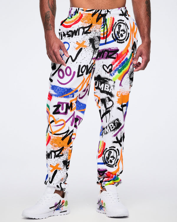Male model wearing colorful graffiti-style pants with white Zumba shoes on a white background.