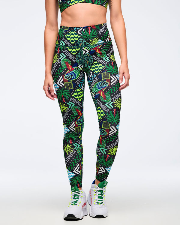 High-waisted Zumba leggings with bold tropical patchwork print featuring parrots, palm leaves, wave graphics, and “ZUMBA” text in green, teal, black, and red. Female model wears the leggings with a matching printed sports bra and white Zumba high-top sneakers on a light gray studio background.