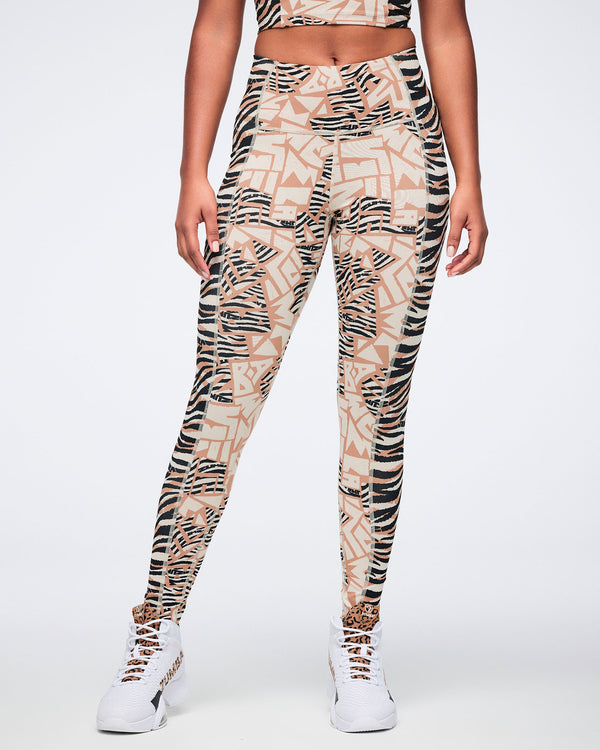 Front view of beige and black mixed-print high-waisted leggings with a fitted full-length silhouette, combining zebra stripes and abstract geometric panels. Female model wears the leggings with white high-top Zumba sneakers and a matching beige and black crop top.