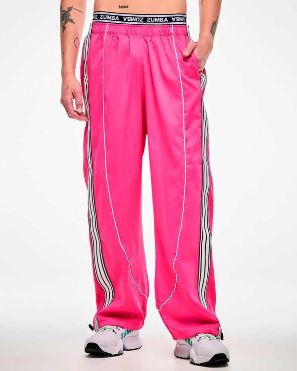 Female model wearing pink track pants with a white and black side stripe and white sneakers, hand in the pocket.
