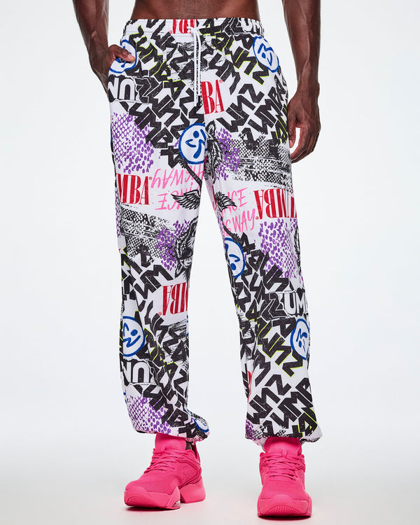 A male model with one hand in his pocket of the white woven pants with an all-over ripped newspaper like pattern with black, white and red ZUMBA text and pink Dance this way text along with other Zumba rock graphics, Zumbitos and purple and black snakeskin patterns, paired with pink shoes.