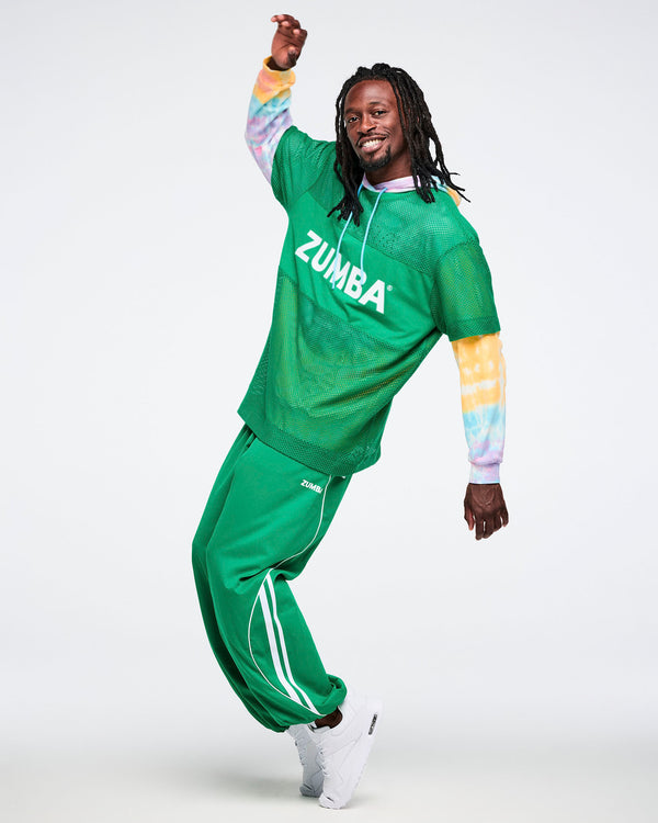 Male model on his toes dancing while wearing a green Zumba mesh top, a tie dye hoodie underneath and green curvy track pants.