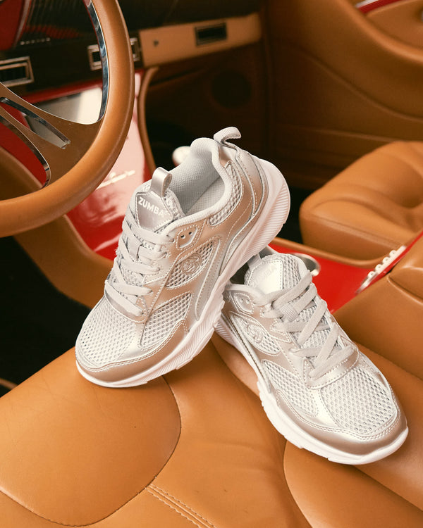 Metallic silver Zumba athletic sneakers with silver mesh panels and white soles. The pair is displayed on a tan leather car seat inside a red vintage car, showing the lace-up design, cushioned shape, and side logo detail.