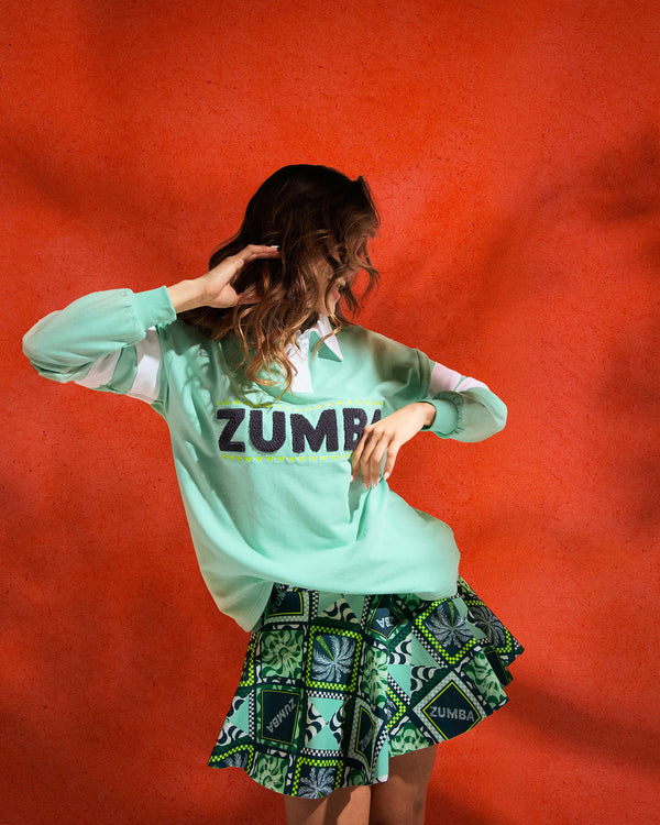 Green Zumba patchwork skort with flared hem and matching printed set peeking underneath. Female model wears a mint pullover sweatshirt with bold “ZUMBA” lettering, posing against a textured red wall.