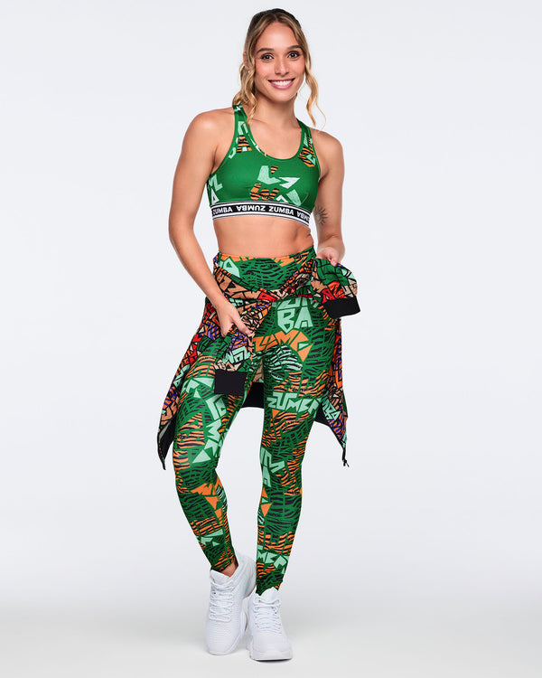 Female model wearing a matching set with a green printed sports bra, high-waisted leggings in coordinating green abstract print, a matching jacket tied at the waist, and white high-top sneakers.
