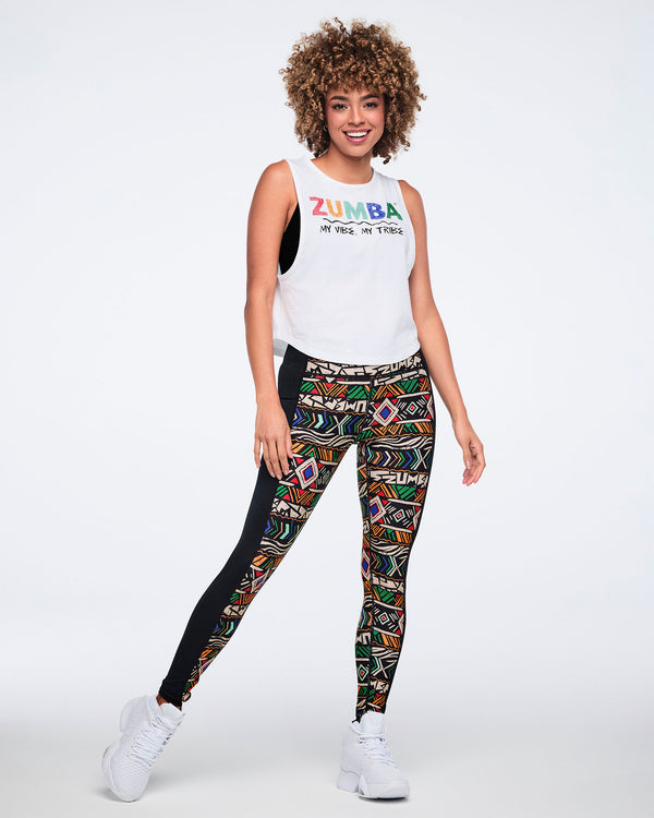 Female model wearing a white sleeveless tank with multicolor Zumba text and “My Vibe, My Tribe” printed on the front, paired with black leggings featuring a bold multicolor geometric print and white high-top sneakers.