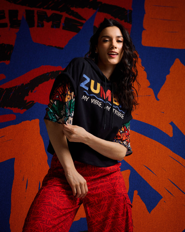 Cropped black graphic hoodie layered on top of a multicolor top on a female model with red printed cargo pants against a bold orange and blue backdrop.