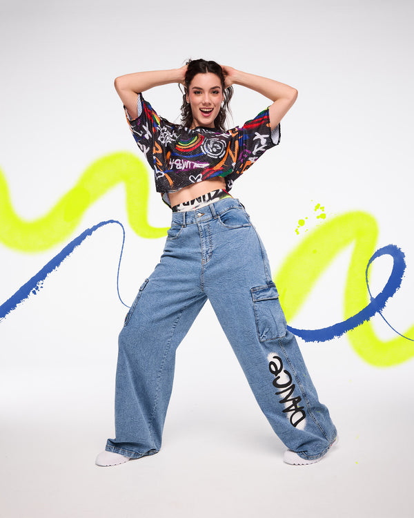 Female model wearing a colorful mesh crop top with denim cargo pants with on a white background with graffiti designs.