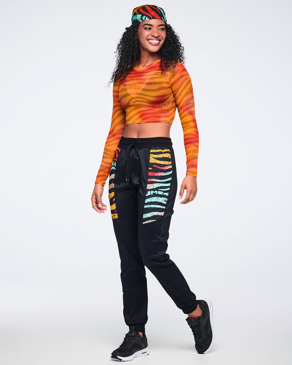 Orange and red zebra-print sheer long-sleeve crop top. Female model wears the top with black jogger pants featuring multicolor side graphics, a matching printed bandana, and black athletic sneakers.