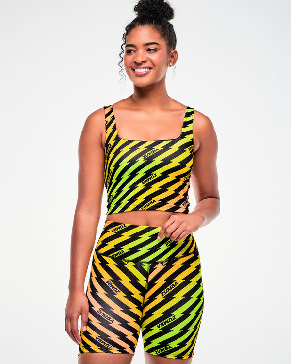 Female model viewed from the front, wearing a yellow, green, and orange ombre crop top and matching biker shorts with a black lightning bolt zigzag print.