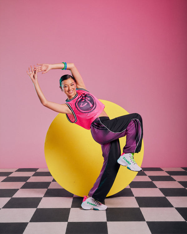 Female model wearing a pink mesh tank with a dot pattern Zumbito logo, paired with black pants with a pink dot side and back panel and white sneakers, against an '80s style backdrop with a checkerboard floor, yellow circle, and red background.