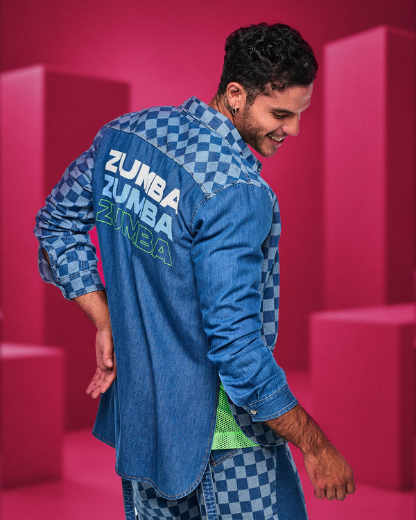 Shown from the back, a male model in a denim shirt with a checkerboard yoke and sleeves and 'ZUMBA' printed three times in white, light blue, and neon green outline, against a pink backdrop.