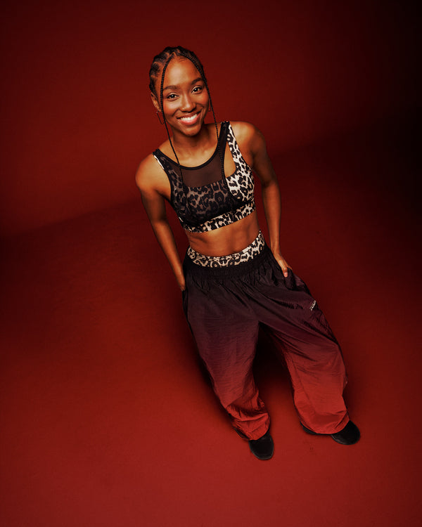 A female model posing in a tan leopard print sports bra with asymmetrical mesh panel and gradient red and black pants, against a solid dark red studio background.