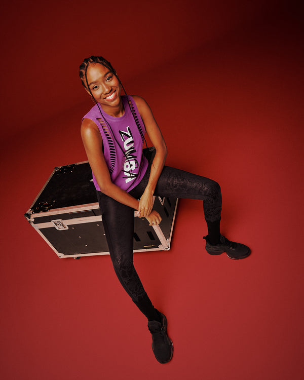 A female model sits on a black equipment case against a solid red studio background. She is wearing a purple tank with cut slashes along sides of front body, with 'ZUMBA' text vertically in white and black, paired with black leggings and black shoes.