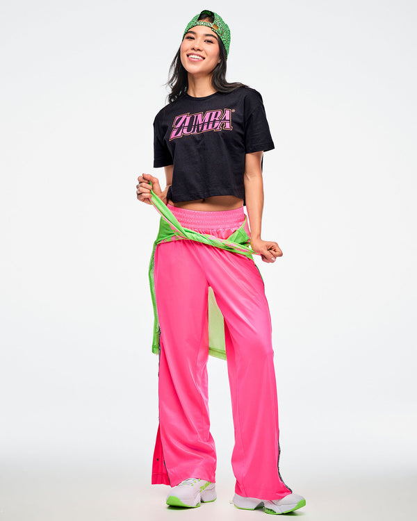 Female model wearing a backward green Zumba hat, a black Zumba T-shirt with pink “ZUMBA” across the chest, pink Zumba pants, and green-and-white shoes, with a green shirt tied around her waist.