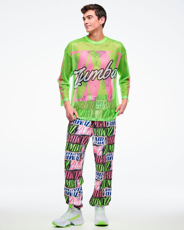Male model wearing a neon green mesh long-sleeve Zumba top with bold pink graphics and white cursive “Zumba,” paired with multicolored Zumba pants and green-and-white shoes.
