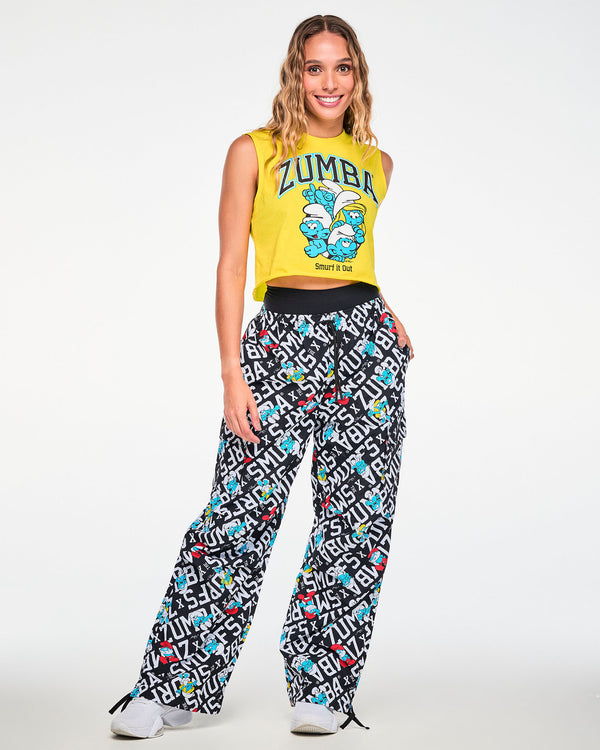 Female model in the yellow crop tank with arched 'ZUMBA' text, huddled Smurfs, and 'Smurf it Out' text, paired with black and white Smurfs print cargo pants and white shoes.