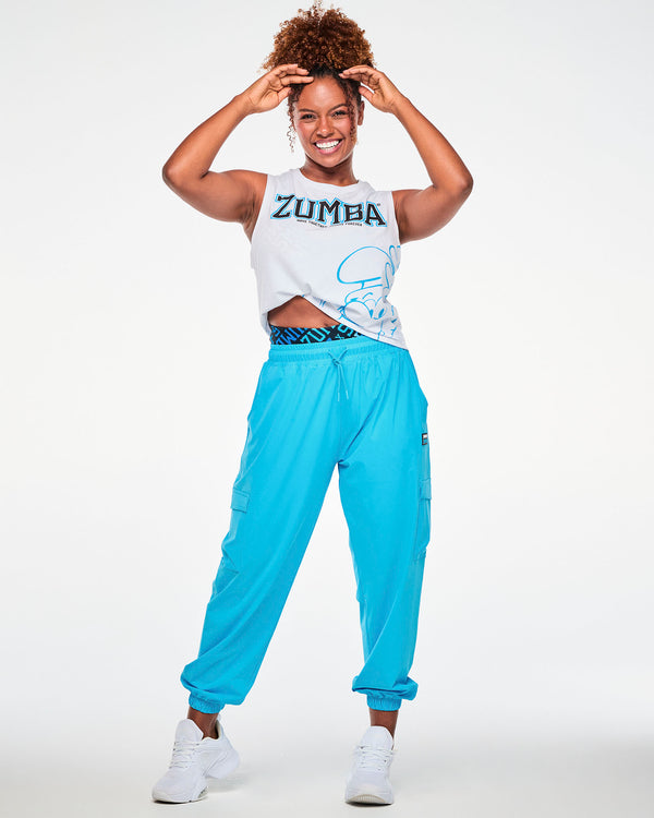Female model with both hands on her head, wearing the white Smurfs graphic muscle tank tied up at the side, paired with aqua blue cargo pants.