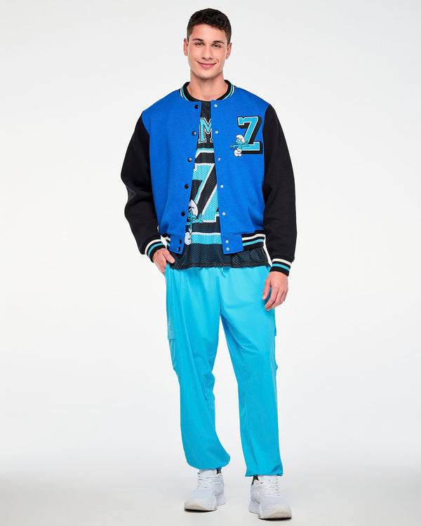 Male model with one hand in his pocket, wearing the blue and black varsity jacket with the chenille 'Z' and Smurf on the chest over a black 'ZUMBA' X 'THE SMURFS' mesh jersey tee, paired with aqua blue cargo pants.