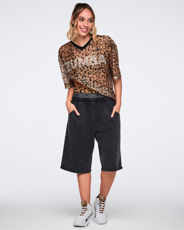 Tan leopard-print mesh top with black abstract spots, a black V-neck collar, oversized fit, and elbow-length sleeves, shown on a smiling female model wearing black acid-wash long shorts and white high-top sneakers with leopard-print details.
