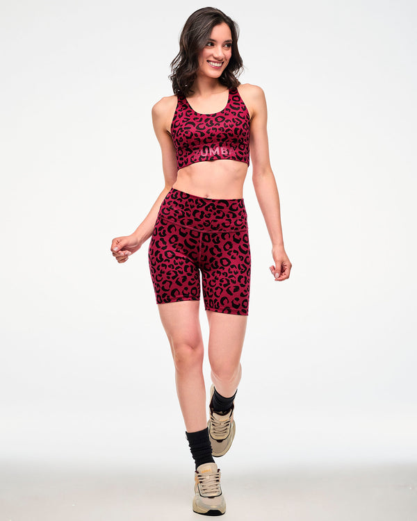 Full view of a female model walking in a dark red longline racerback bra with a black leopard print and red 'ZUMBA' text on the underband, paired with matching biker shorts, black socks, and beige shoes.