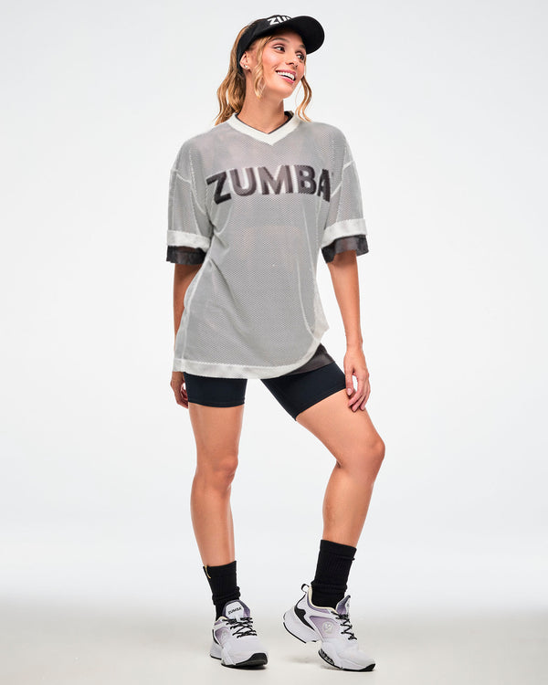 Female model in the white mesh oversized top styled with a black 'ZUMBA' visor, black biker shorts and socks and white shoes.