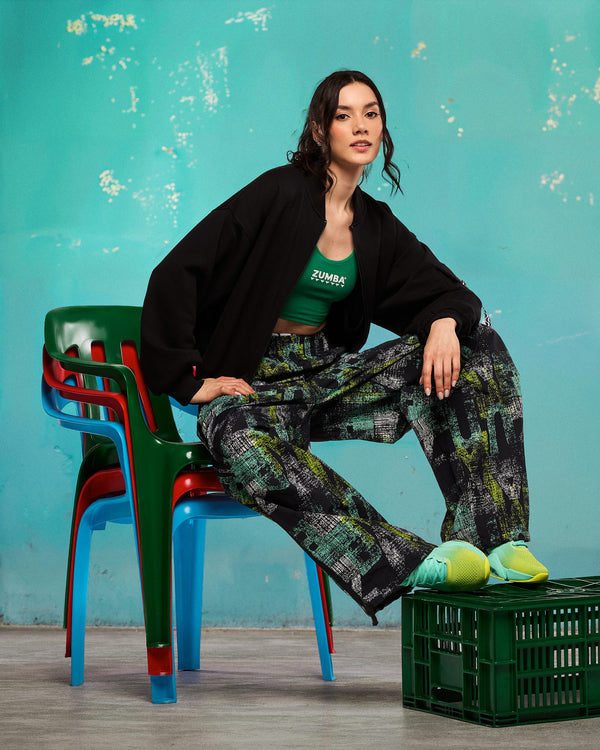 Lifestyle shot of a female model seated on stacked chairs wearing the black wide-leg printed track pants, green tank, oversized black jacket, and aqua-to-lime sneakers.