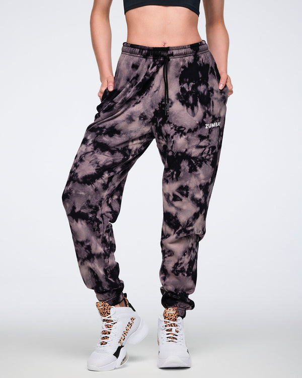 Black and tan tie-dye sweatpants with a small white Zumba logo on the front. Female model wears the relaxed-fit joggers with a drawstring waistband, cuffed ankles, a black sports bra, and white high-top sneakers with black and tan accents.