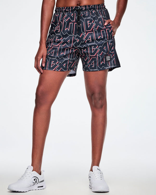 Female model with a hand in her left pocket, wearing mesh shorts featuring a black, white, and red ZUMBA text pattern, paired with white shoes.