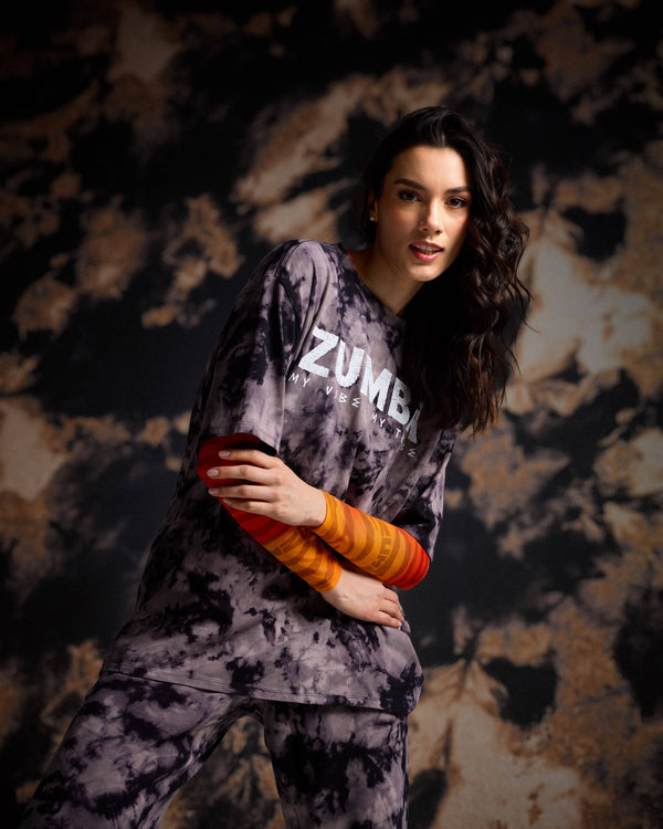 Female model in a black and tan tie-dye Zumba tee, layered on a top with orange striped arm sleeves, matching pants, and before a dark cloudy background.