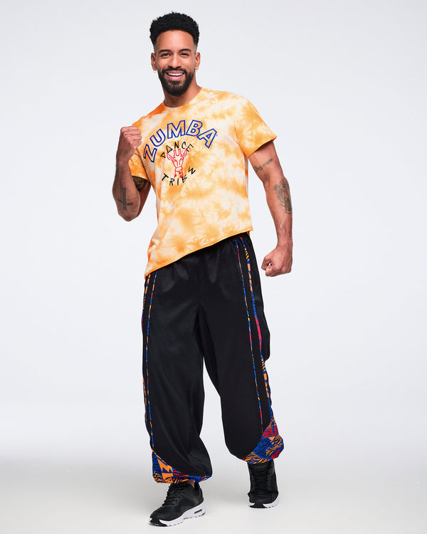 Male model wearing an oversized orange and white tie-dye T-shirt with blue Zumba text and a “Dance Tribe” giraffe graphic, paired with black wide-leg athletic pants with multicolor printed trim and black sneakers.