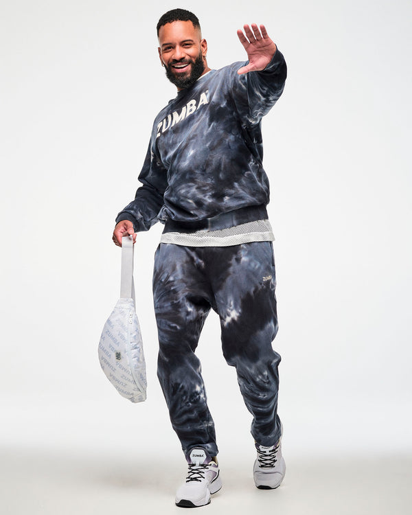 Black and grey tie-dye crewneck sweatshirt with cream 'ZUMBA' text, paired with matching tie-dye joggers and a white 'ZUMBA'-print crossbody bag, male model.