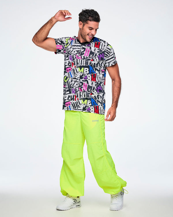Full view of a male model dancing while wearing a multi-colored ZUMBA graphic tee, paired with yellow pants and white shoes.