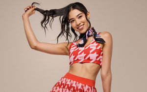 Zumba red-and-pink printed sports bra worn by female model playing with her hair, in a matching pleated skort, styled for Zumba workouts, and vibrant athleisure wear.