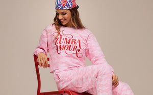 Pink Zumba Wear “Zumba Amour” sweatshirt and jogger set with heart pattern, modeled by a woman seated on a red chair, showcasing women’s dance-inspired athleisure apparel.
