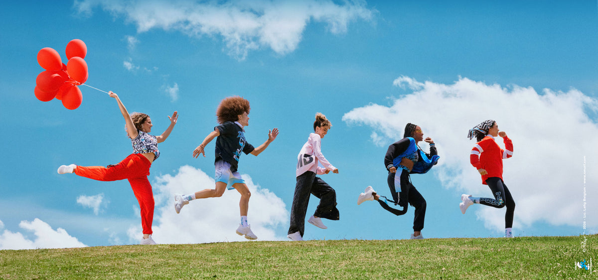 Five male and female models leap and run in a line across a grassy hilltop under a bright blue sky with white clouds, the lead model holding a bunch of red balloons. Outfits include red cargo pants, a red and white striped button-up, a blue and black varsity jacket, a red rugby polo, and Smurfs logo prints. A small Smurfs copyright credit runs along the right edge.