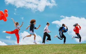 Small banner with five male and female models running and leaping in a line across a grassy hilltop under a bright blue sky with white clouds, the lead model trailing red balloons. Their Zumba X The Smurfs outfits include red cargo pants, a black mesh jersey with ombre shorts, a striped button-up, a varsity jacket, and a red rugby polo.
