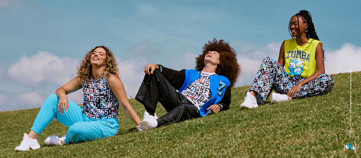 Three models relax on a grassy hillside under a soft blue sky: a female model in a black and white Smurfs print tank with aqua blue pants, a male model in a blue and black varsity jacket over a Smurfs print tee with black pants, and a female model in a yellow 'ZUMBA' Smurfs tank with Smurfs print pants. A small Smurfs copyright credit runs along the right edge.