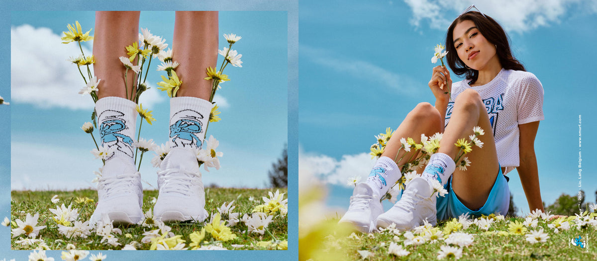 Two side-by-side photos. On the left, inside a soft blue border, a close-up of legs in white Smurf crew socks and white shoes with daisies tucked in, standing in flower-strewn grass. On the right, a female model sits among daisies in a white mesh 'ZUMBA' tee, light blue shorts, and the Smurf socks, holding a flower under a blue sky. A small Smurfs copyright credit runs along the right edge.