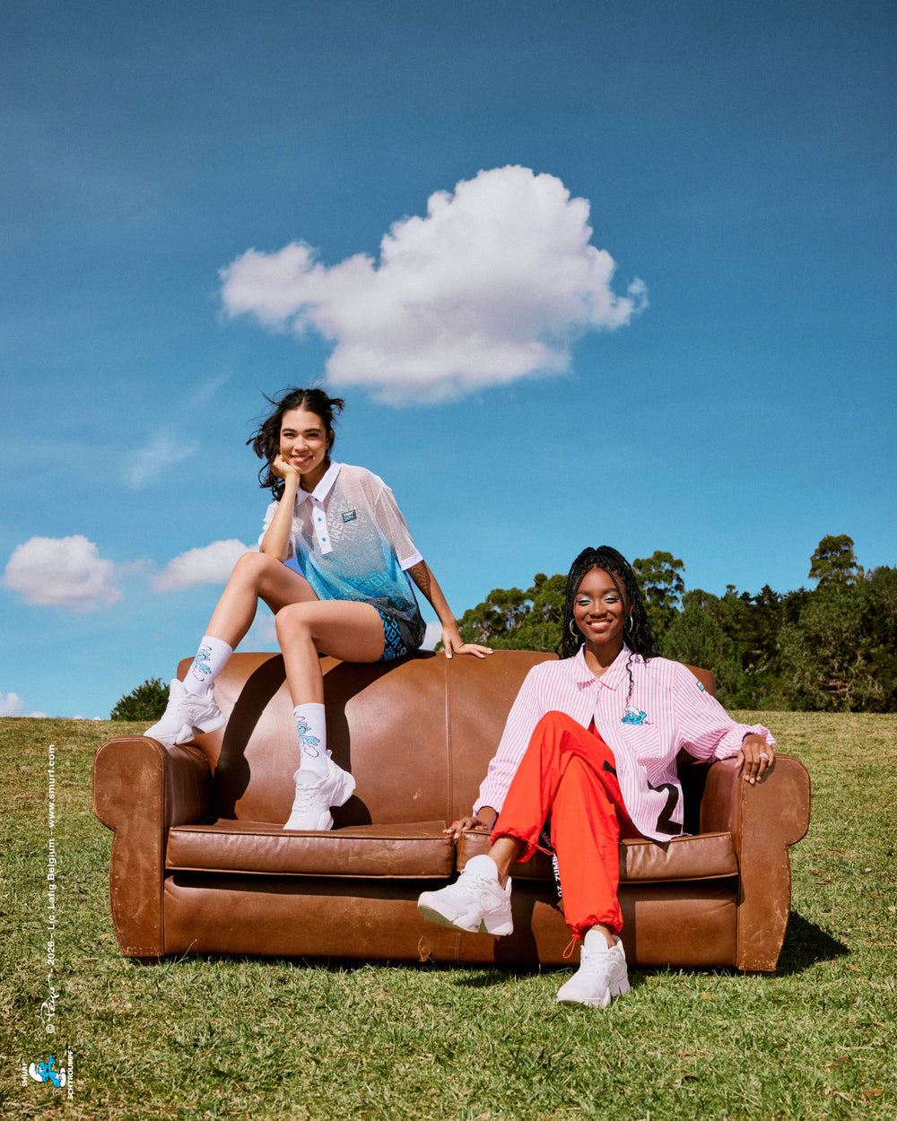 Two female models on a brown leather couch on a grassy hill under a blue sky with a single white cloud: one perched on the backrest in a white-to-blue mesh polo, print shorts, and Smurf socks, the other seated in a red and white striped button-up with red cargo pants. A small Smurfs copyright credit runs along the left edge.