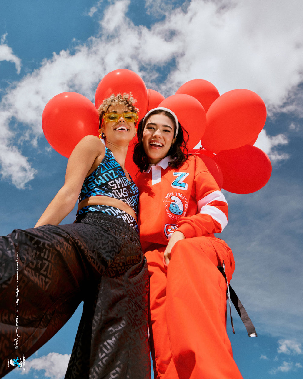 Low-angle shot of two female models in front of a bunch of red balloons against a cloudy blue sky, one in a blue and white 'ZUMBA' X 'THE SMURFS' print bra with black tonal print pants, the other in a red rugby polo with embroidered 'Z' and Smurf patches and red cargo pants. A small Smurfs copyright credit runs along the left edge.