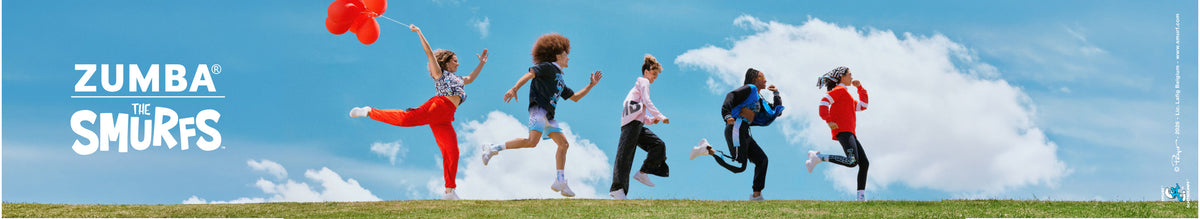 Five male and female models leap and run in a line across a grassy hilltop under a blue sky with white clouds, the lead model holding red balloons. White 'ZUMBA' text over a thin line and 'THE SMURFS' logo sit on the left side of the banner. Outfits include red cargo pants, a red and white striped button-up, a blue and black varsity jacket, and a red rugby polo. A small Smurfs copyright credit runs along the right edge.